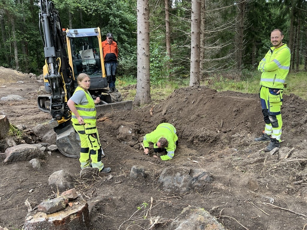 Man ligger på marken och gräver med litet verktyg. Bredvid syns tre personer vid utgrävningen. Bakom utgrävning står en schaktmaskin.