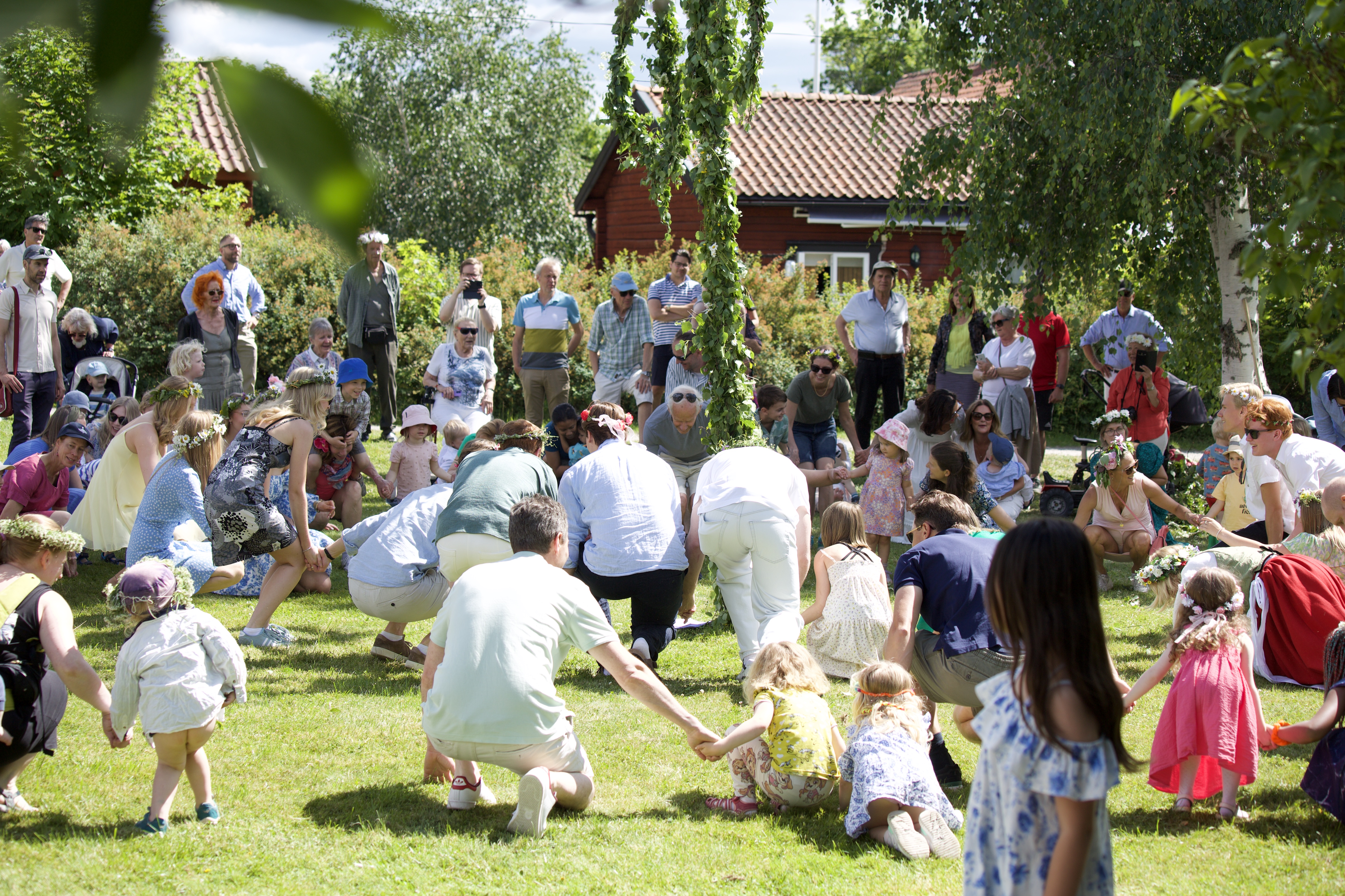 Mycket människor av blandade åldrar som firar midsommar och dansar runt en midsommarstång.