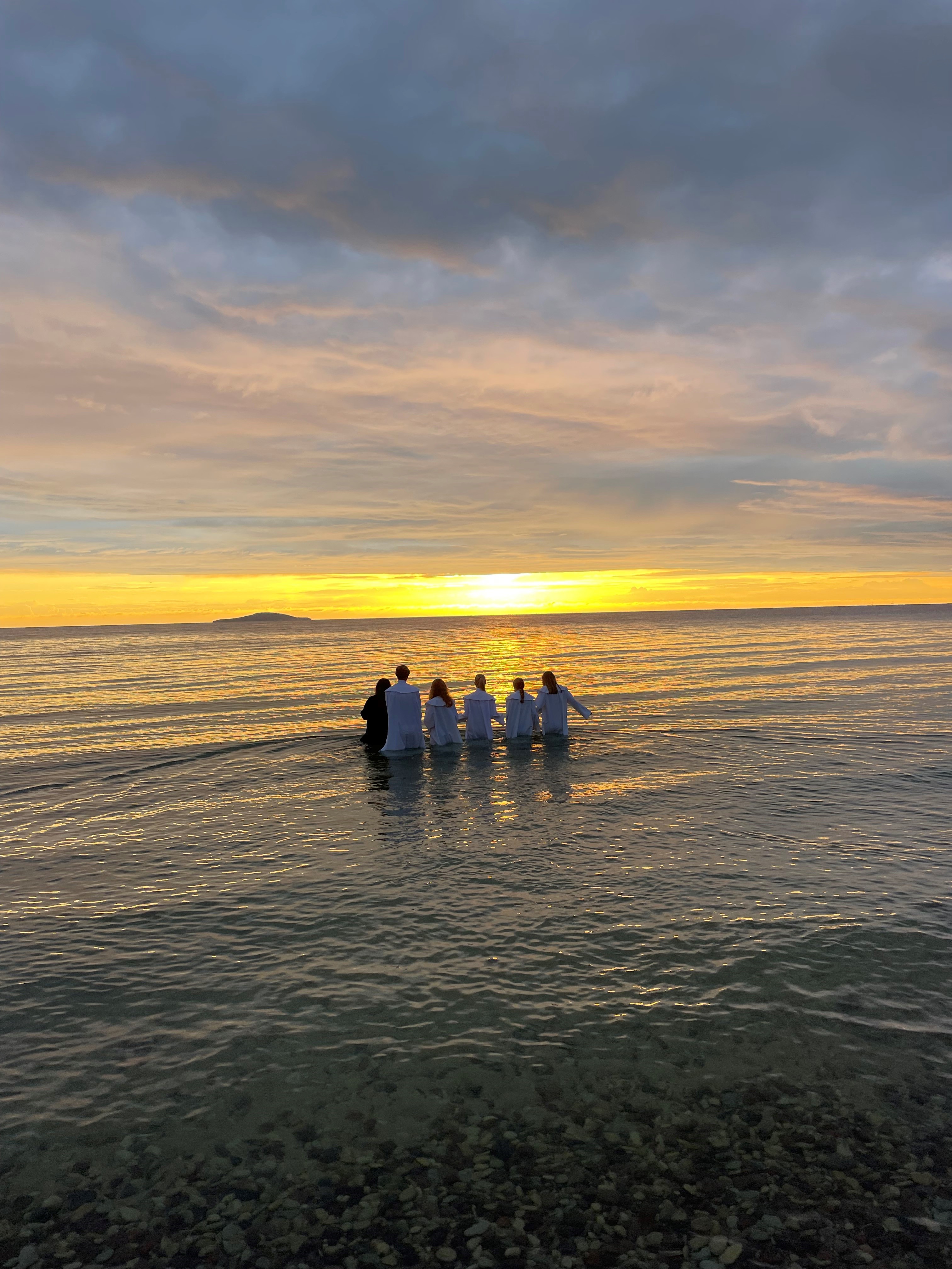 En strand i skymningen, konfirmander står i grupp i vita dräkter.