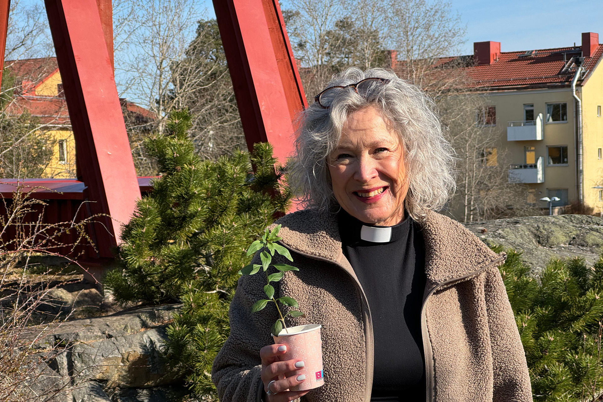 Marie Rehnstam står frmför klockstapeln utanför Årsta kyrka och håller i en liten planta som snart ska planteras ut i våra pallkragar.
