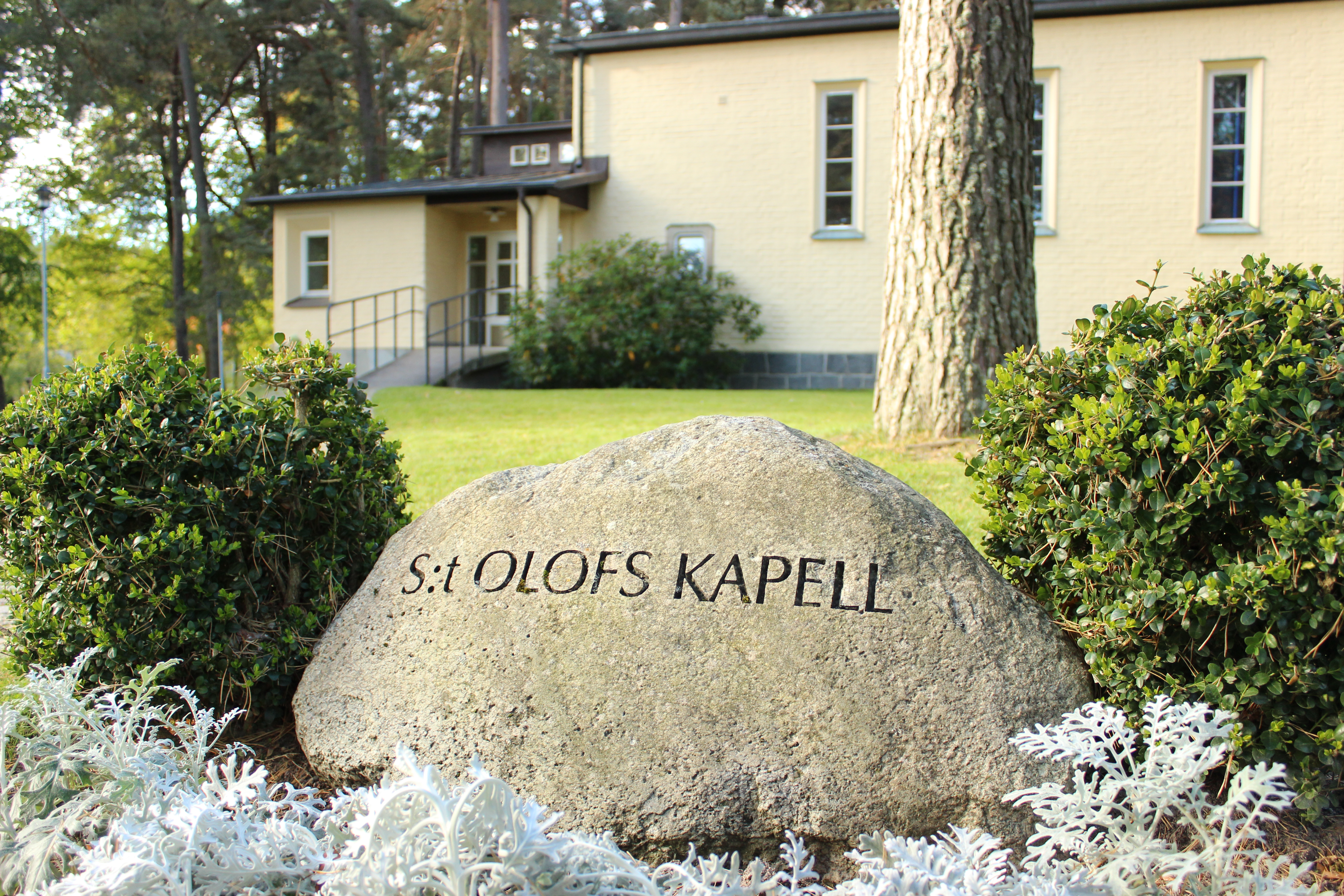 Sten med texten S:t Olofs kapell. I bakgrunden ett ljusgult kapell med ramp till igången.