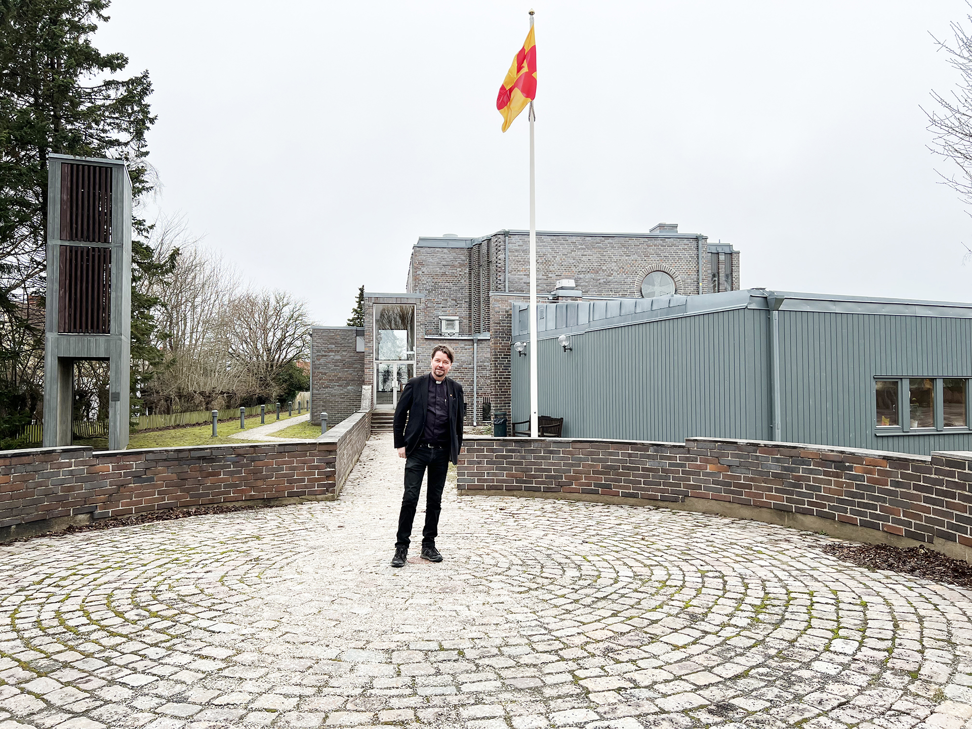 Präst står framför kyrkobyggnad med flagga