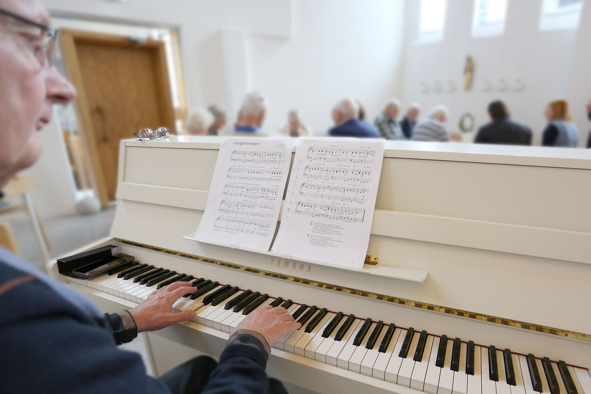 En man spelar på ett vitt piano och andaktsdeltagarna sitter längre bort.