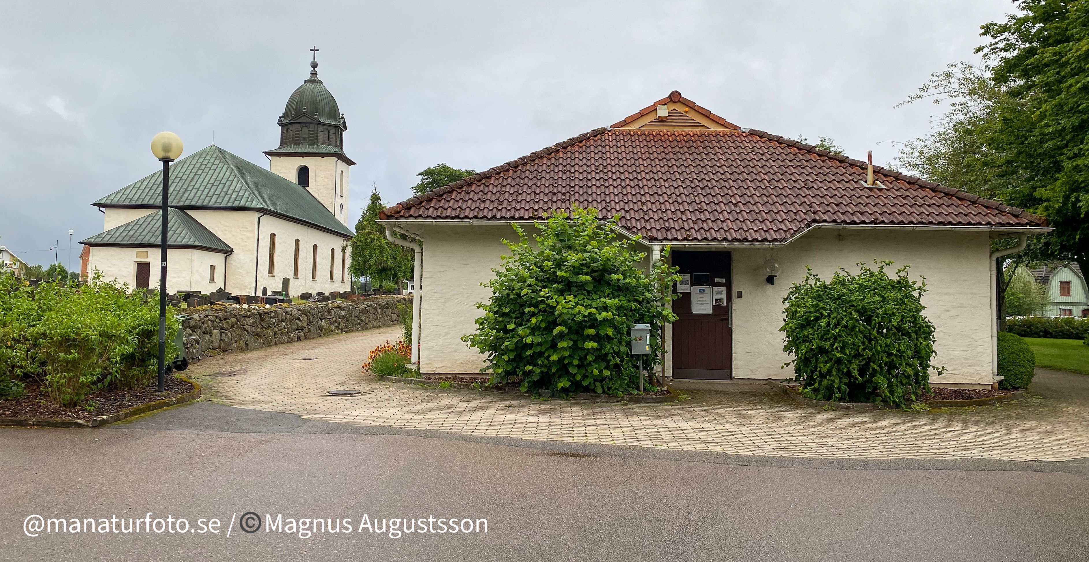 En låg vit byggnad med gröna buskar framför. Bredvid ligger en vit kyrka.