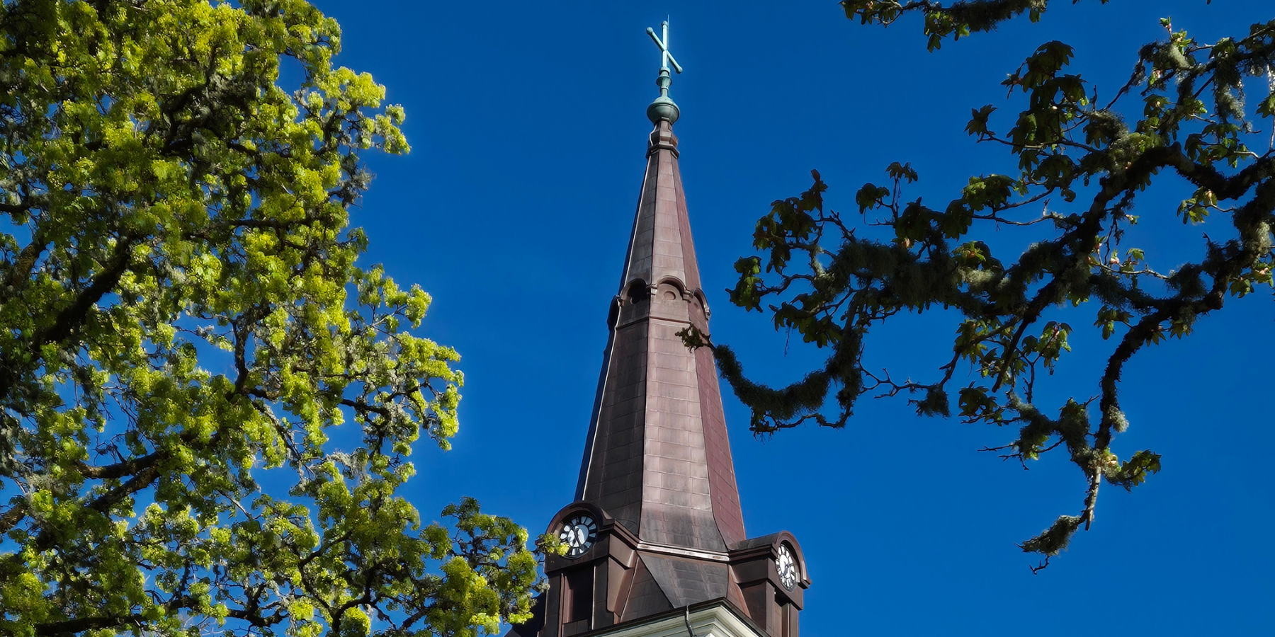Örby kyrkas tornspira mot blå himmel och med träd runt