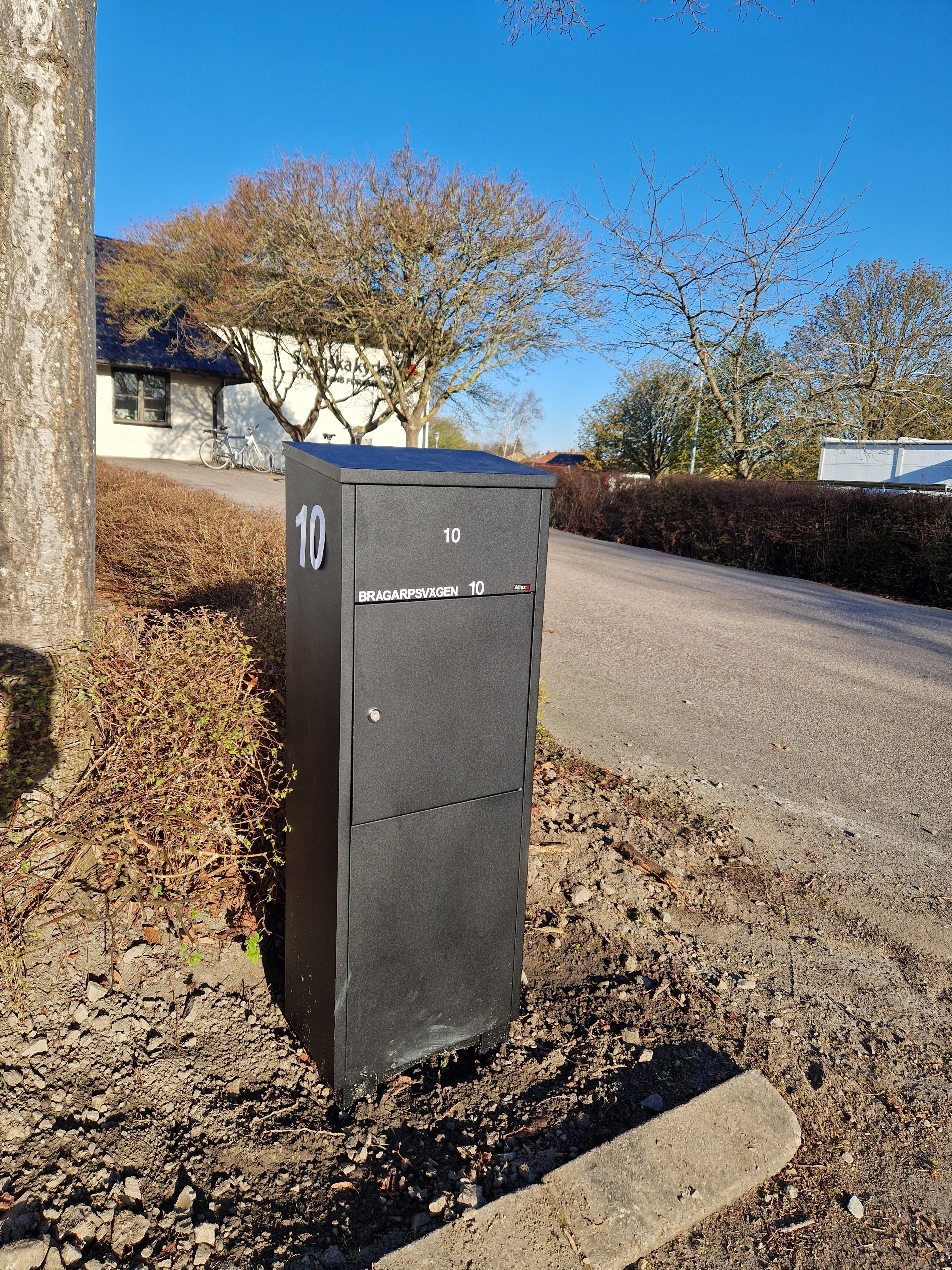 En svart, hög brevlåda med adressen i vit text, vid vägkanten framför församlingsgården.