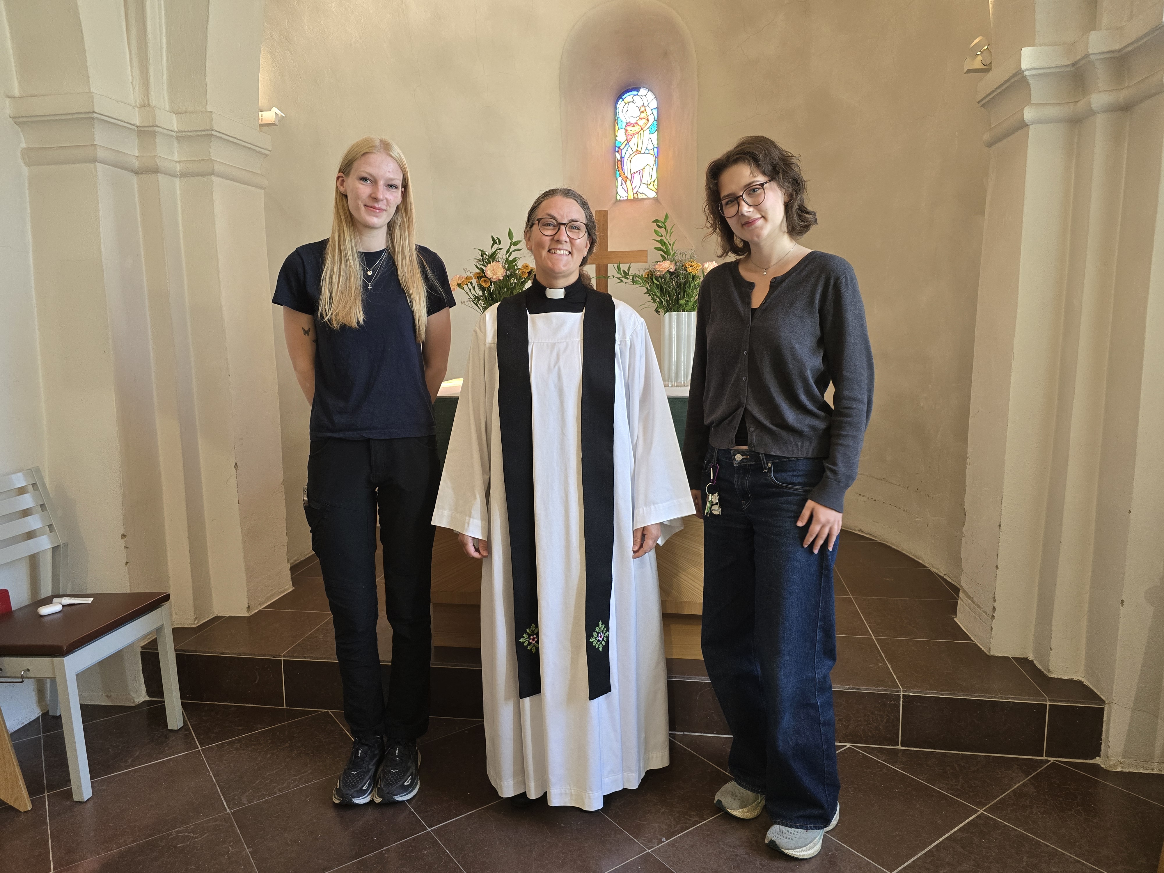 Präst klädd i vitt med svart stola framför altaret i Brågarps kyrka. På vardera sidan står en ung kvinna i svarta kläder.