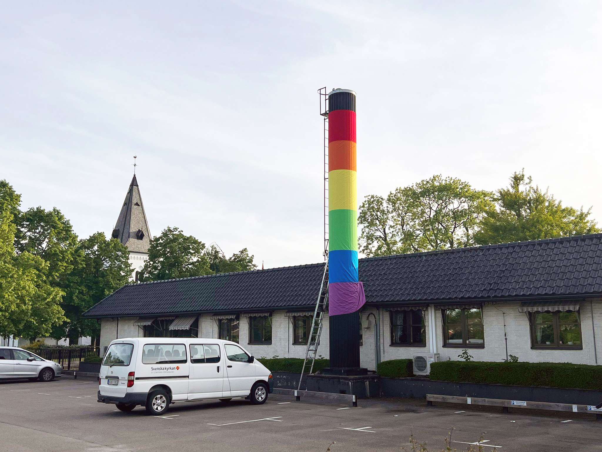 Församlingsgården med Brågarps kyrka i bakgrunden och skorstenen inklädd i breda ränder i regnbågens färger.
