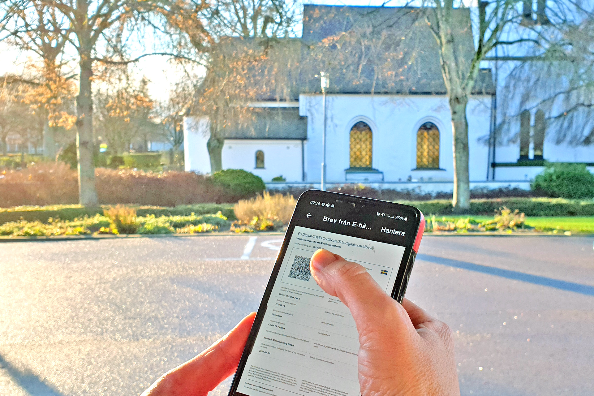 Hand håller mobiltelefon med covidbevis. Brågarps kyrka i bakgrunden.