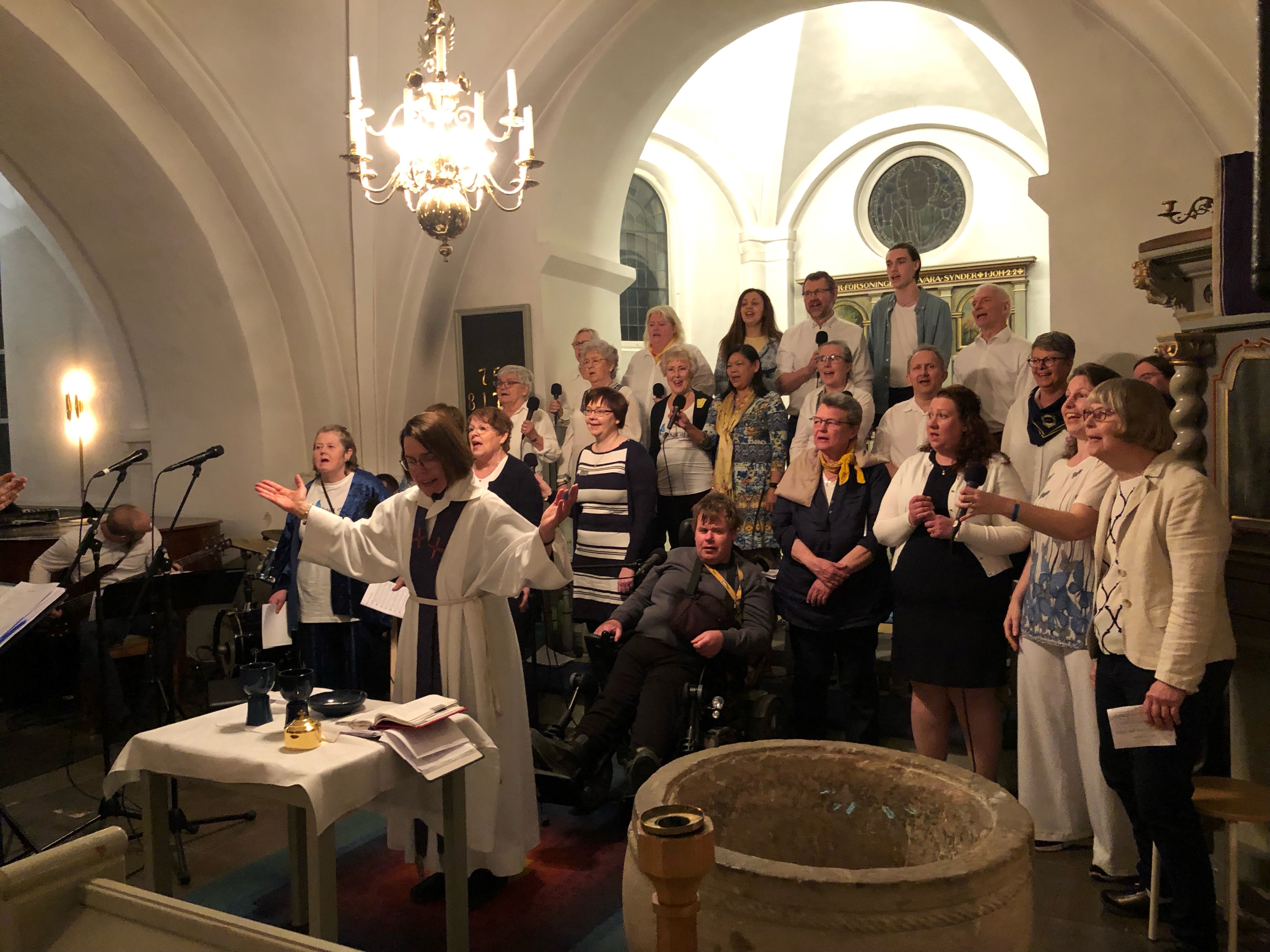 Kören står uppställd i koret i Nevishögs kyrka, bakom prästen Ildiko. Alla sjunger.