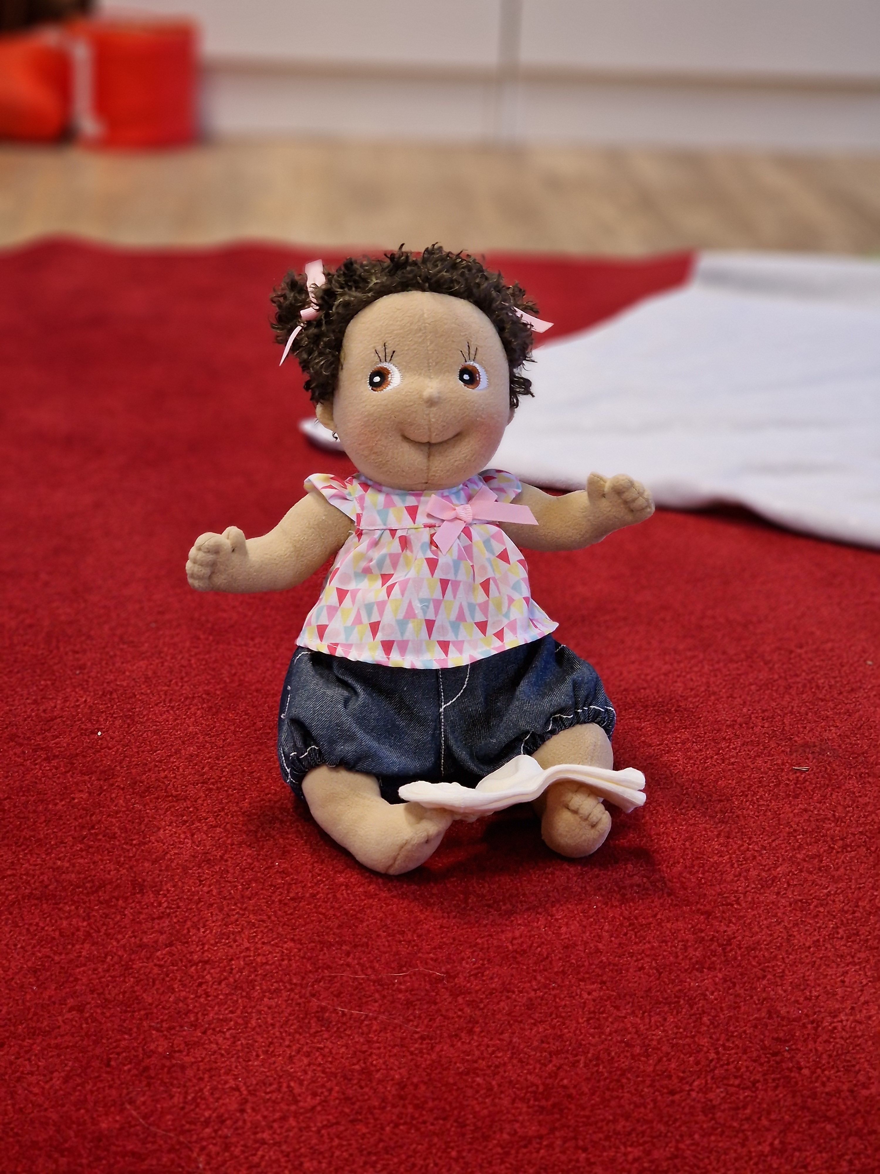 Tyg docka sittandes på röd matt, vit filt i bakgrunden. 