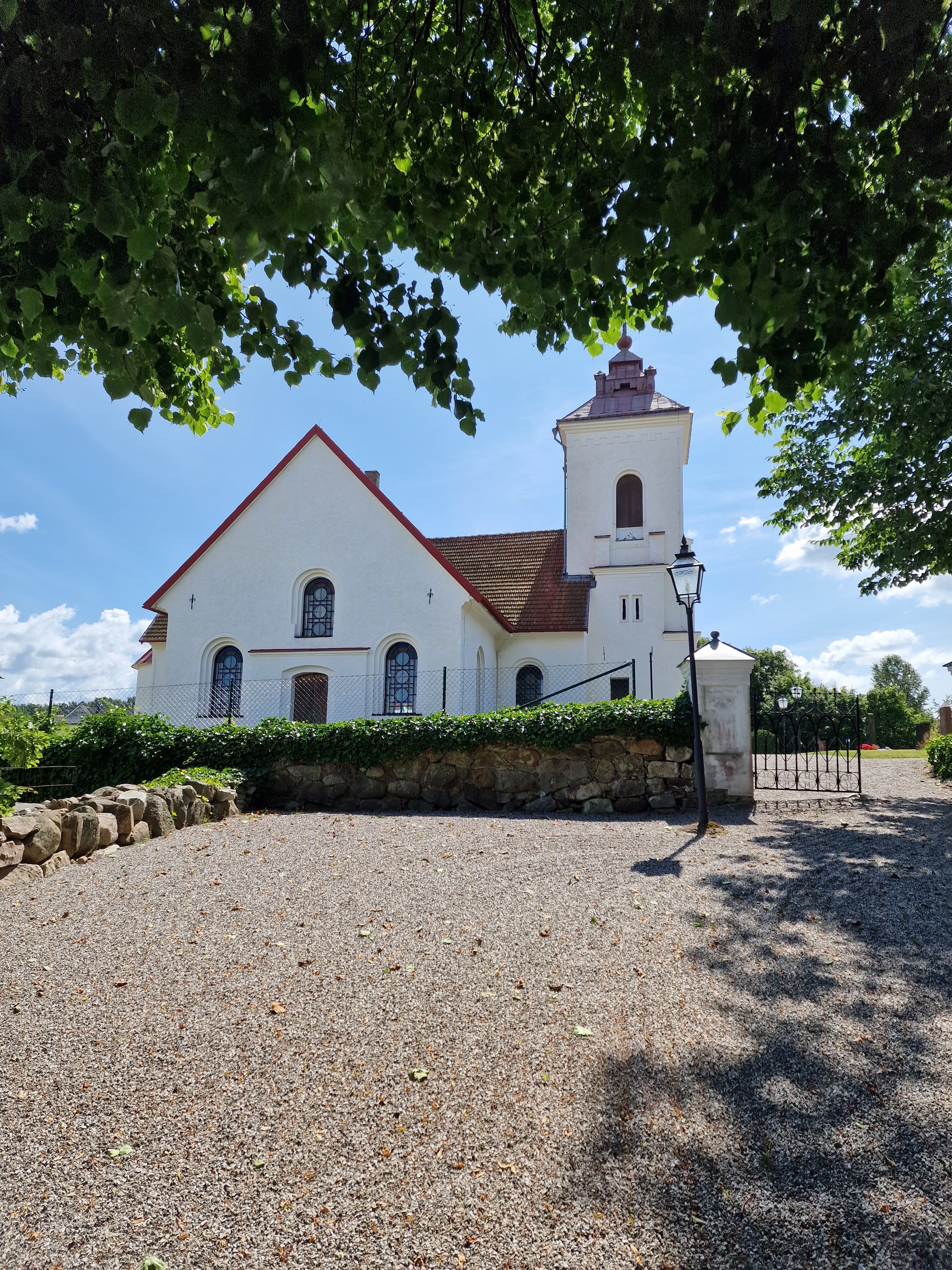 Vit kyrkobyggnad med rött tegeltak med kyrkomuren framför.