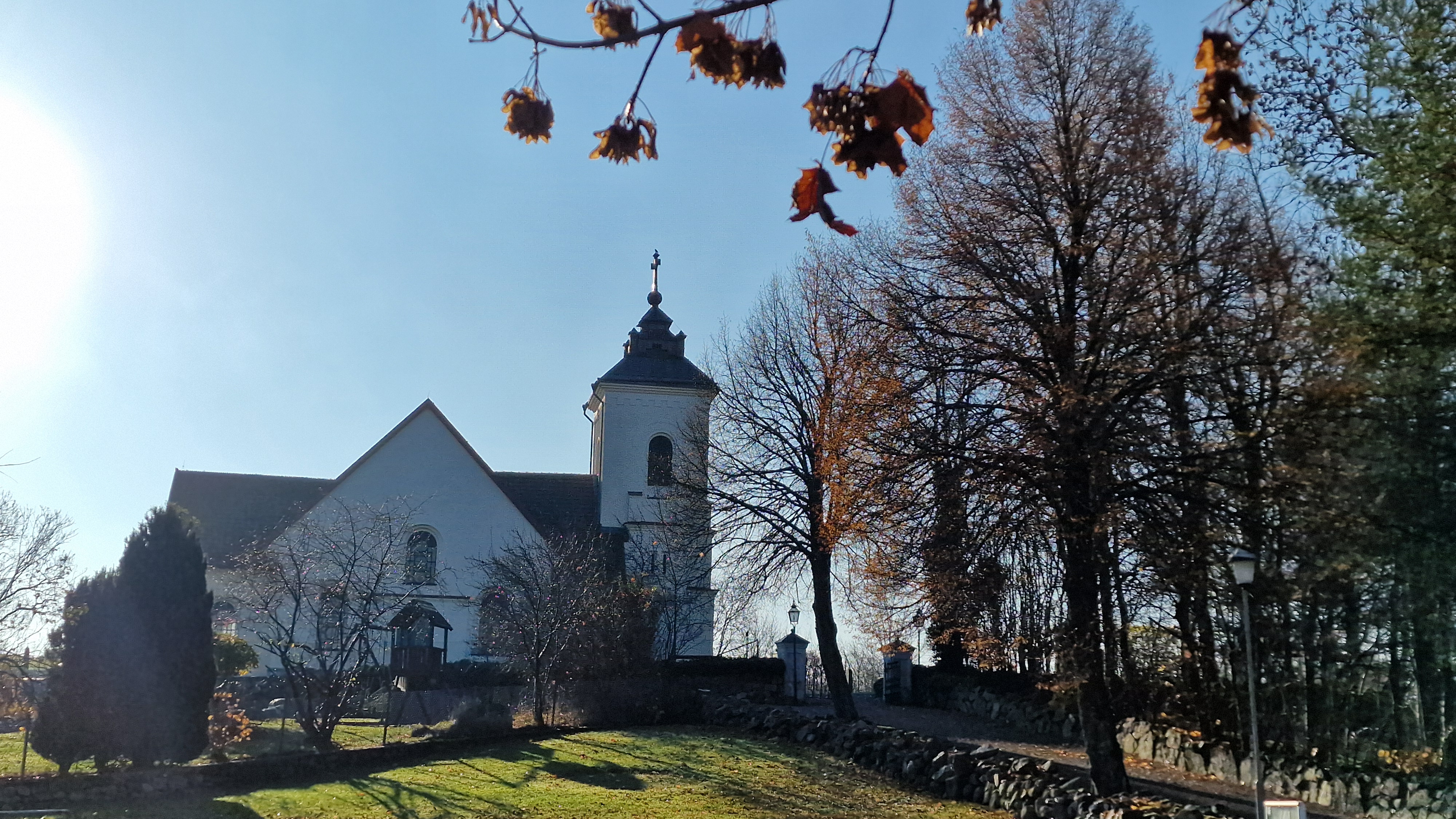 Vit kyrkobygnad uppe på en lägre höjd med träd med fåtal bruna löv kvar