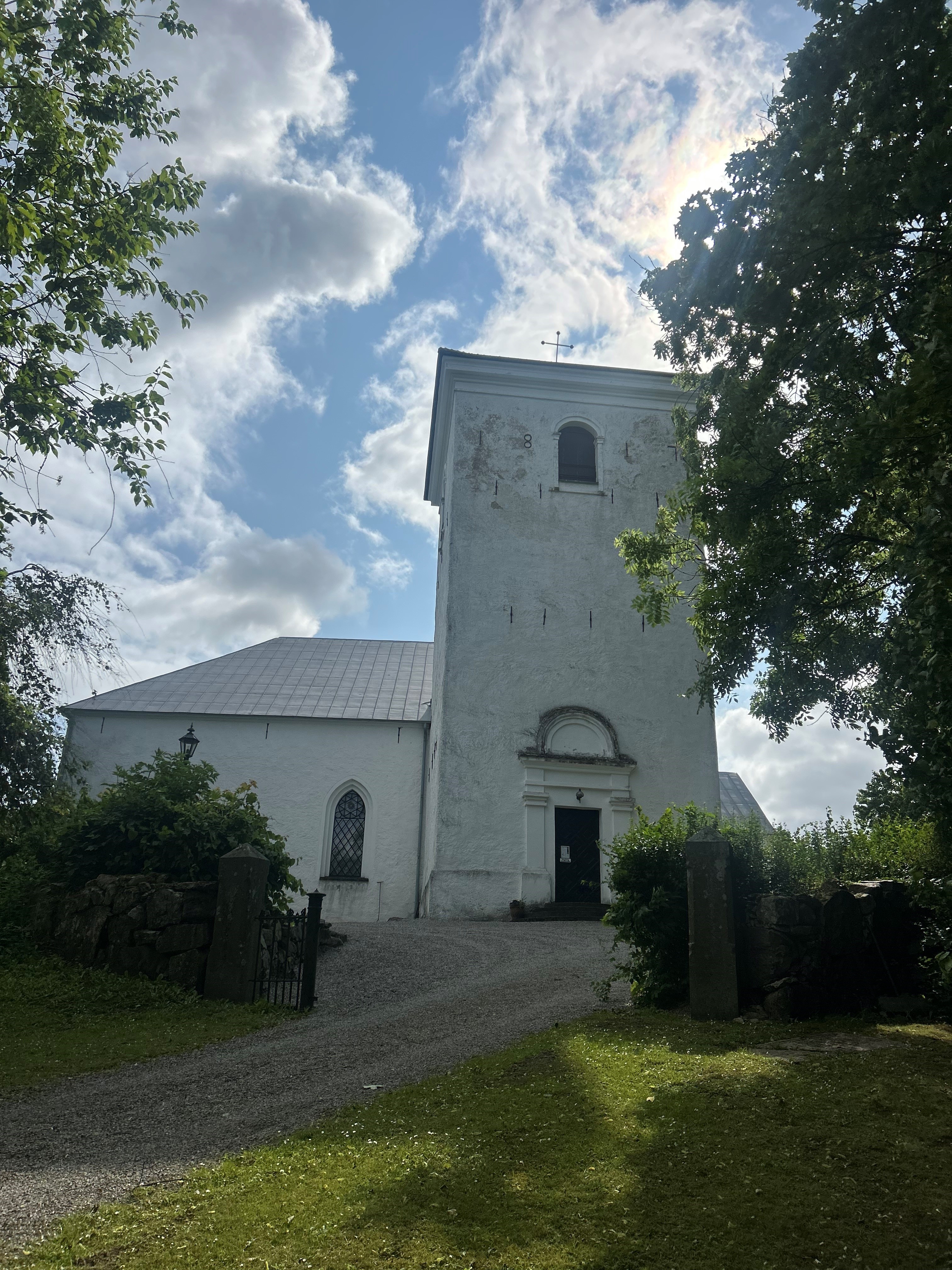 En vit kyrkobyggnad på en backe omringad om grönska. 