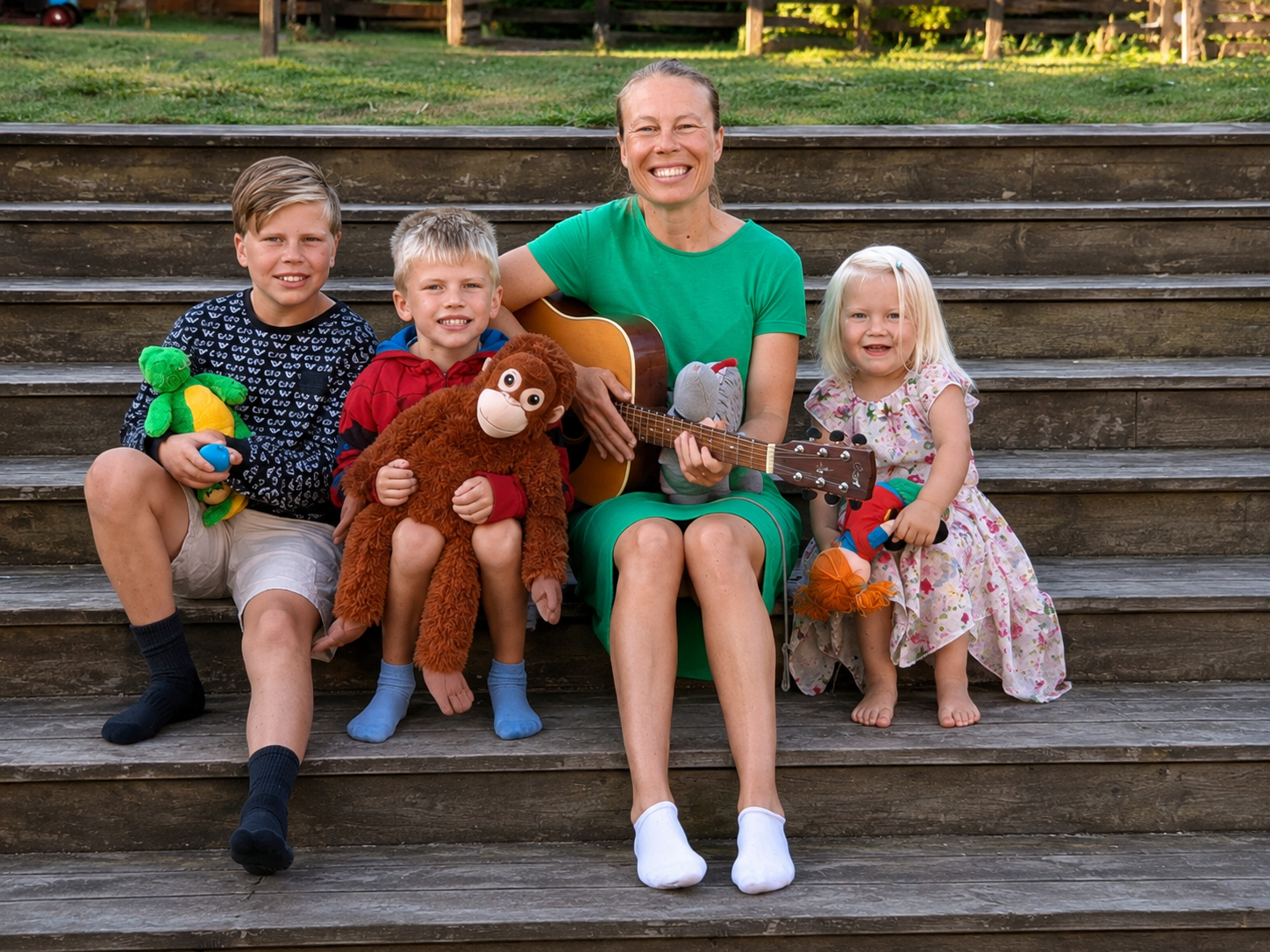 Kvinna med gitarr tillsammans med tre barn i färgglada kläder sittandes på en trappa utomhus.