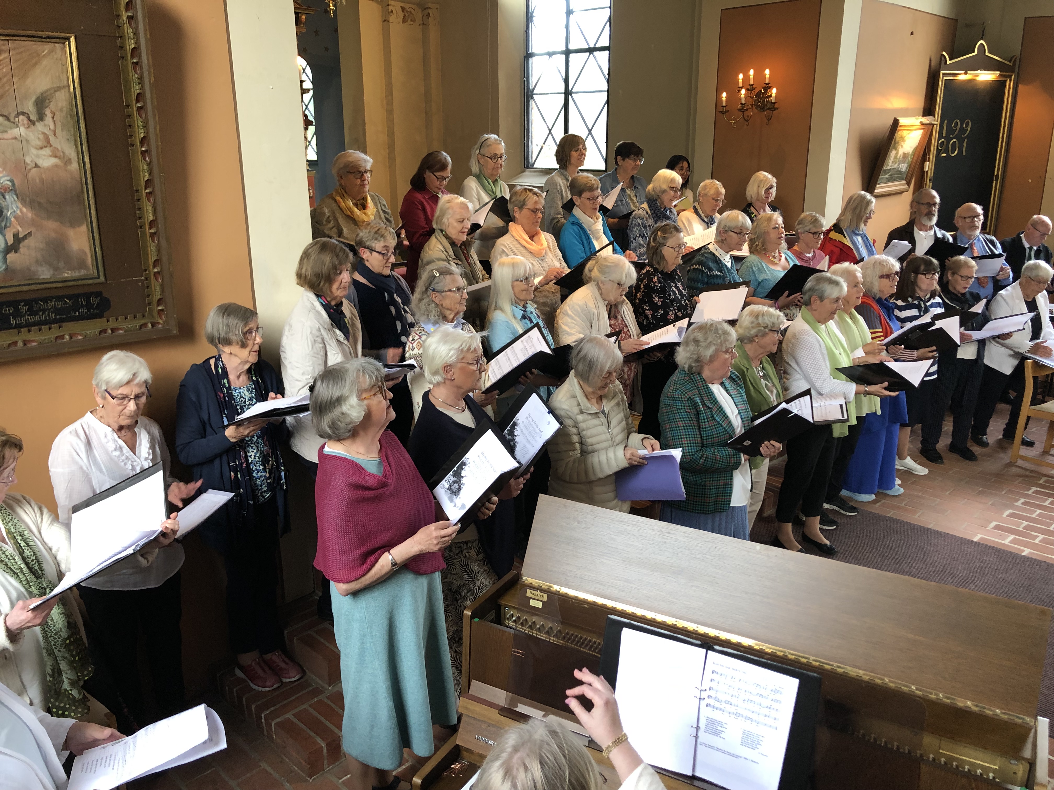 Ett stort körsällskap står tätt tillsammans inne i en kyrka med högt i tak, ljusa väggar och höga fönster. Miljön är varm och stillsam, med dagsljus som faller in genom fönstren.
