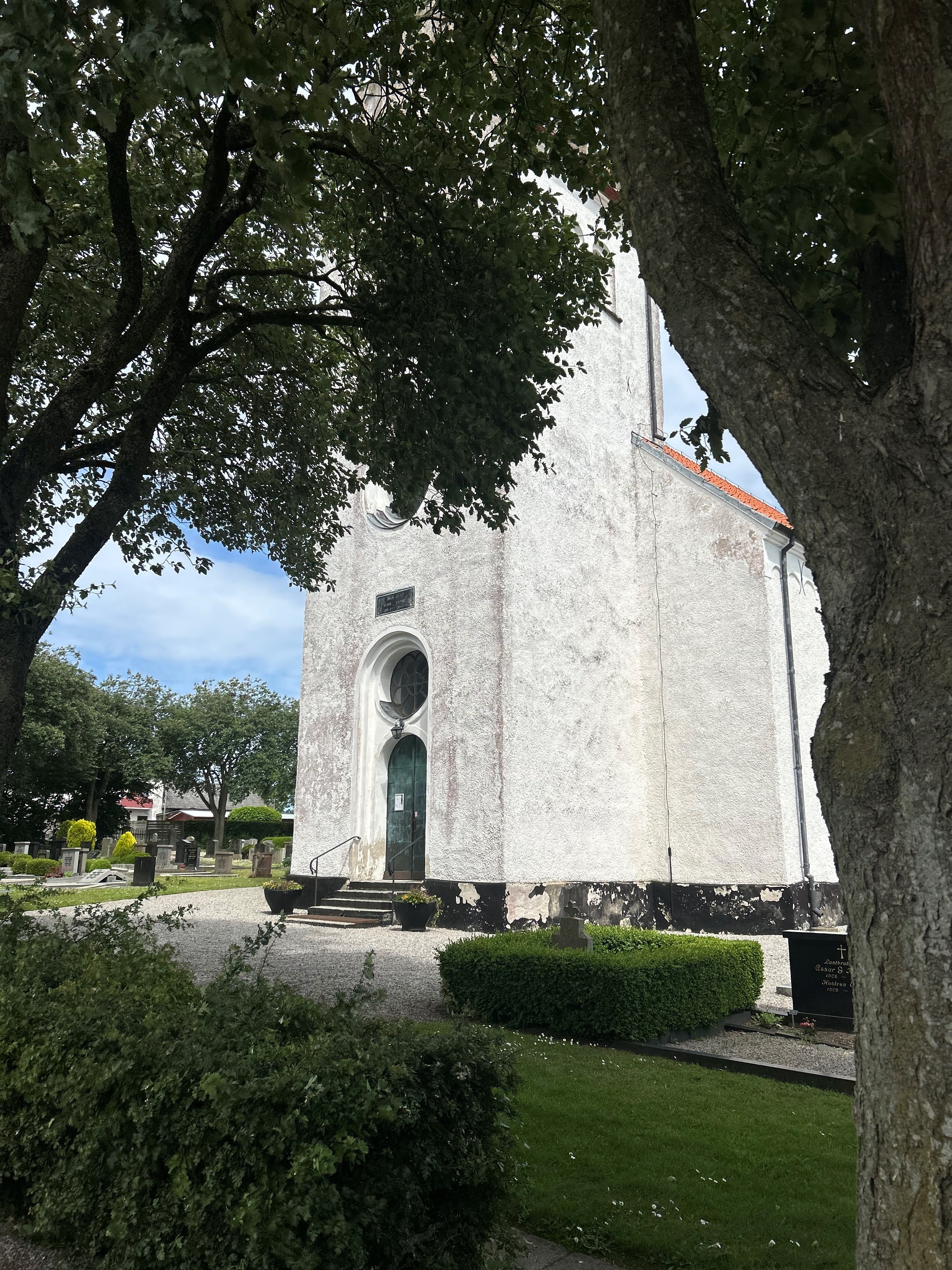 Vit kyrkobyggnad, omringad med skog och buskar.
