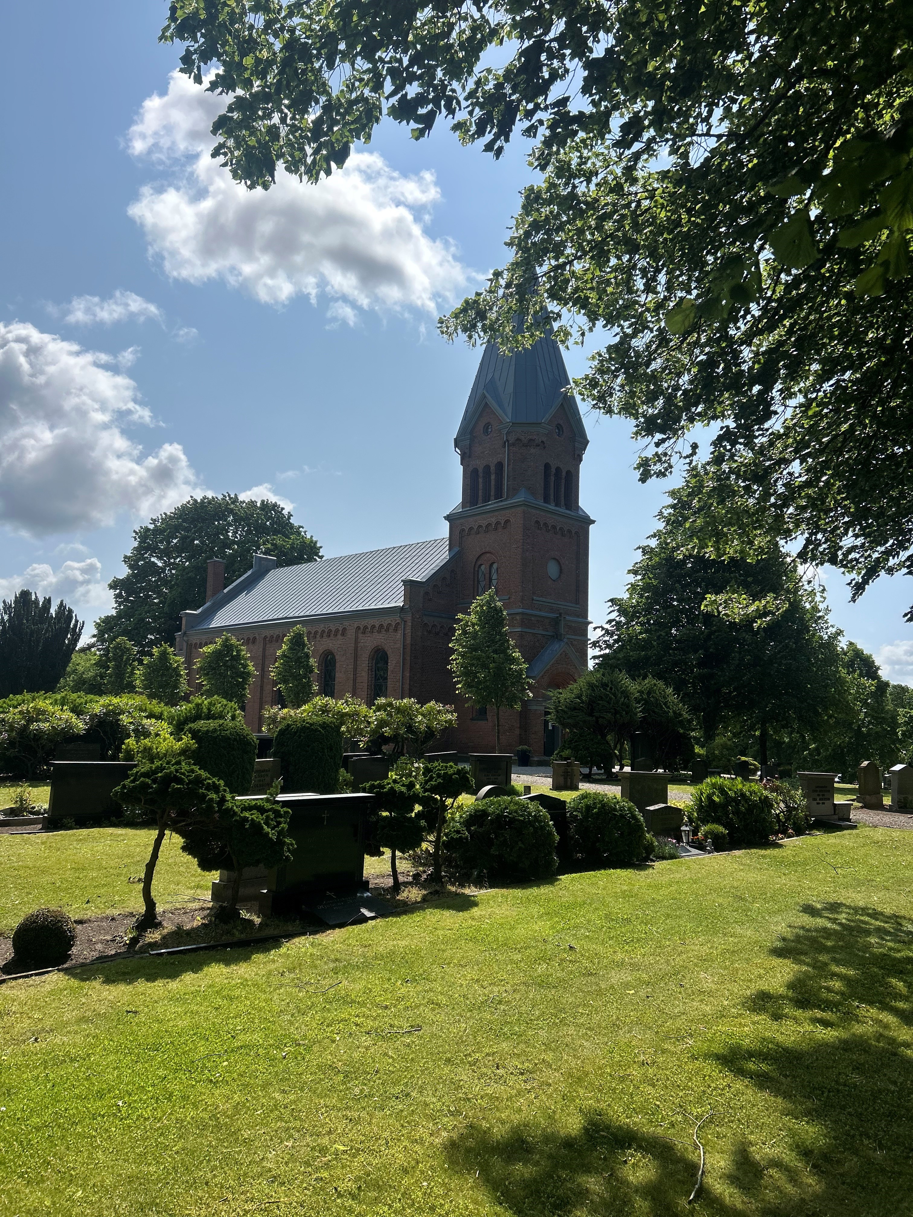 Röd tegelbyggnad omringad av grönska och blå himmel.
