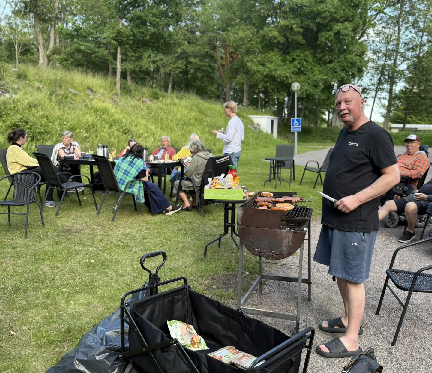 En man står vid en grill, i bakgrunden sitter folk och äter.
