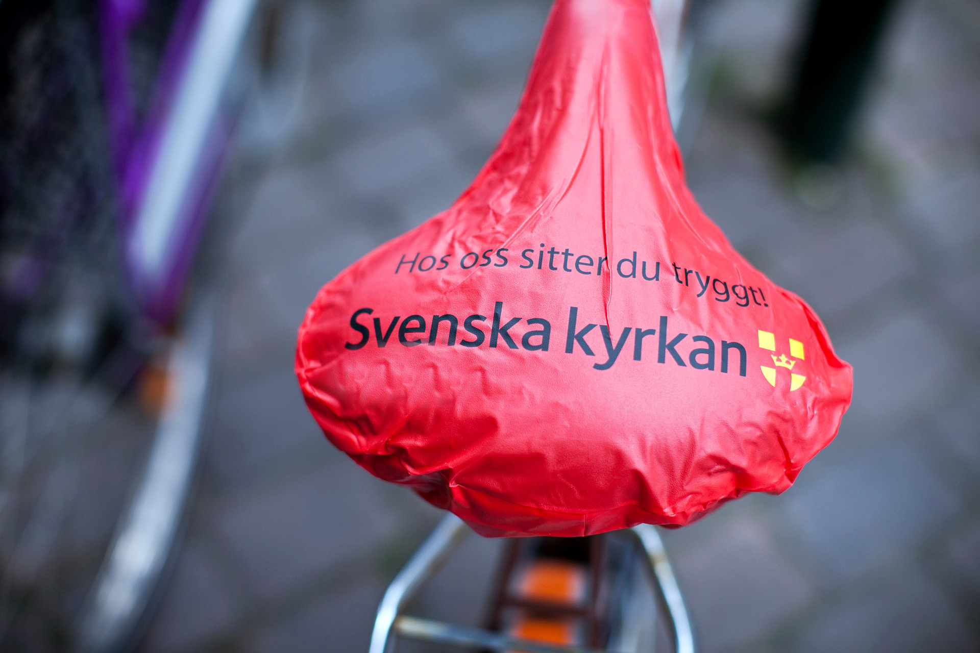 Rött cykelsadelskydd med Svenska kyrkans logo och texten hos oss sitter du säkert.