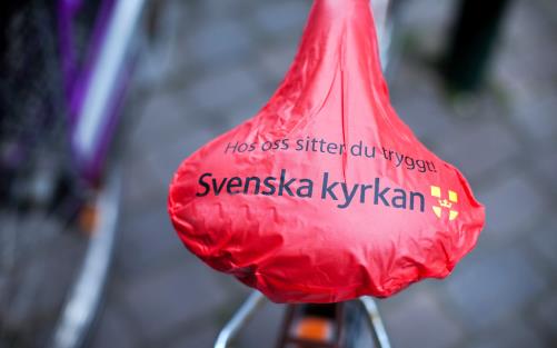 Rött cykelsadelskydd med Svenska kyrkans logo och texten hos oss sitter du säkert.