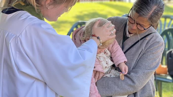 Kvinnlig präst häller vatten på ett barn huvud. Barnet hålls i famnen av sin mormor. Detta är ett utomhusdop.