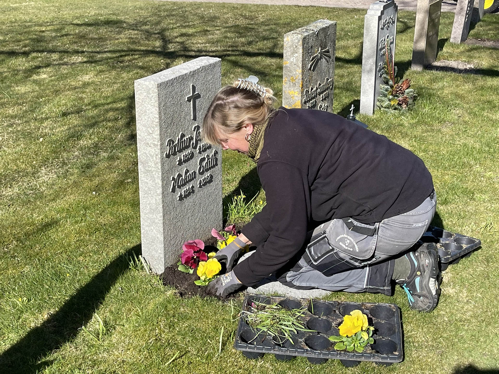 Kvinna ligger på knä framför en gravsten och planterar färgglada blommor.