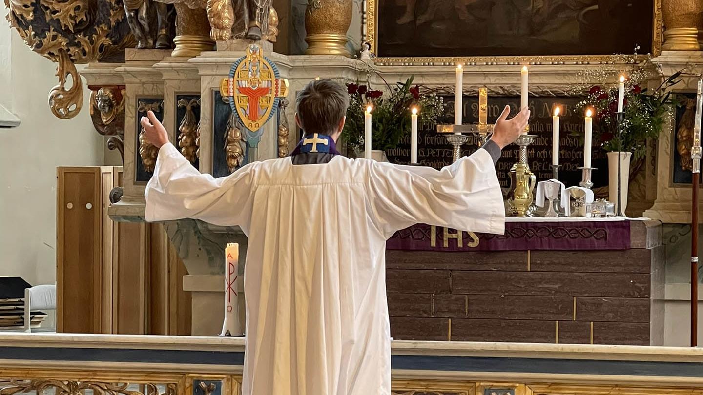 En präst i liturgiska kläder står vänd mot altaret och höjda händer.