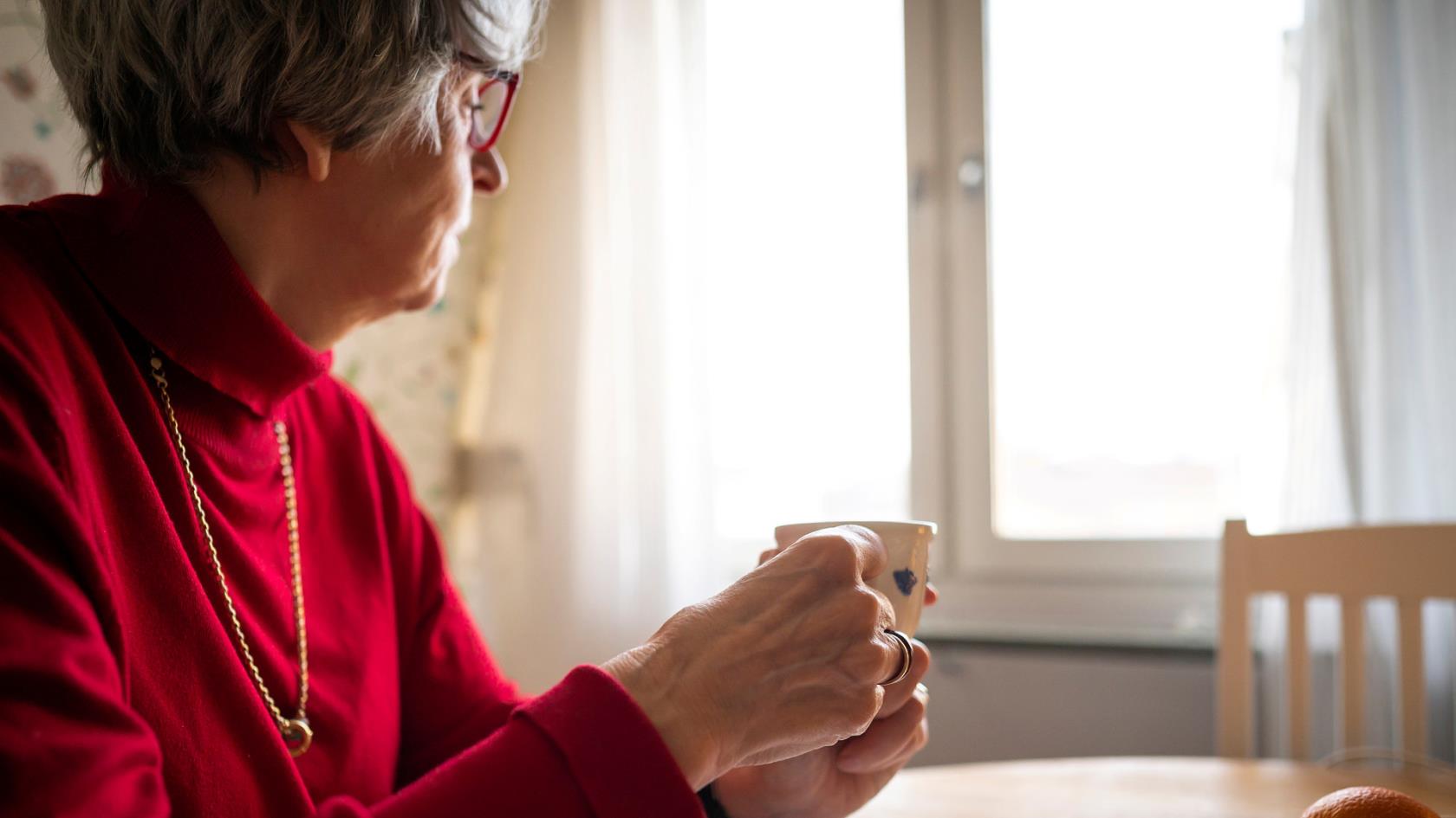Kvinna i röd tröja dricker en kopp kaffe själv vid köksbordet och tittar ut genom fönstret.