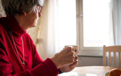 Kvinna i röd tröja dricker en kopp kaffe själv vid köksbordet och tittar ut genom fönstret.