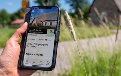 Bilden visar en mobiltelefon och en hand mot en lantlig bakgrund