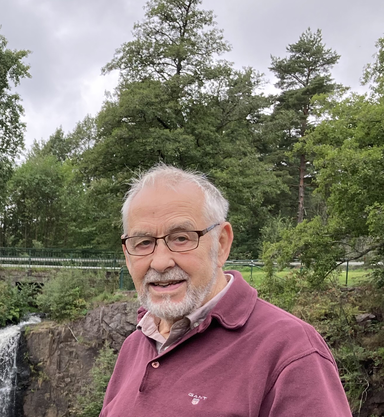 En äldre, leende man med skägg, vitt hår, glasögon och en lila tröja fram ett ett naturområde med träd och buskar.