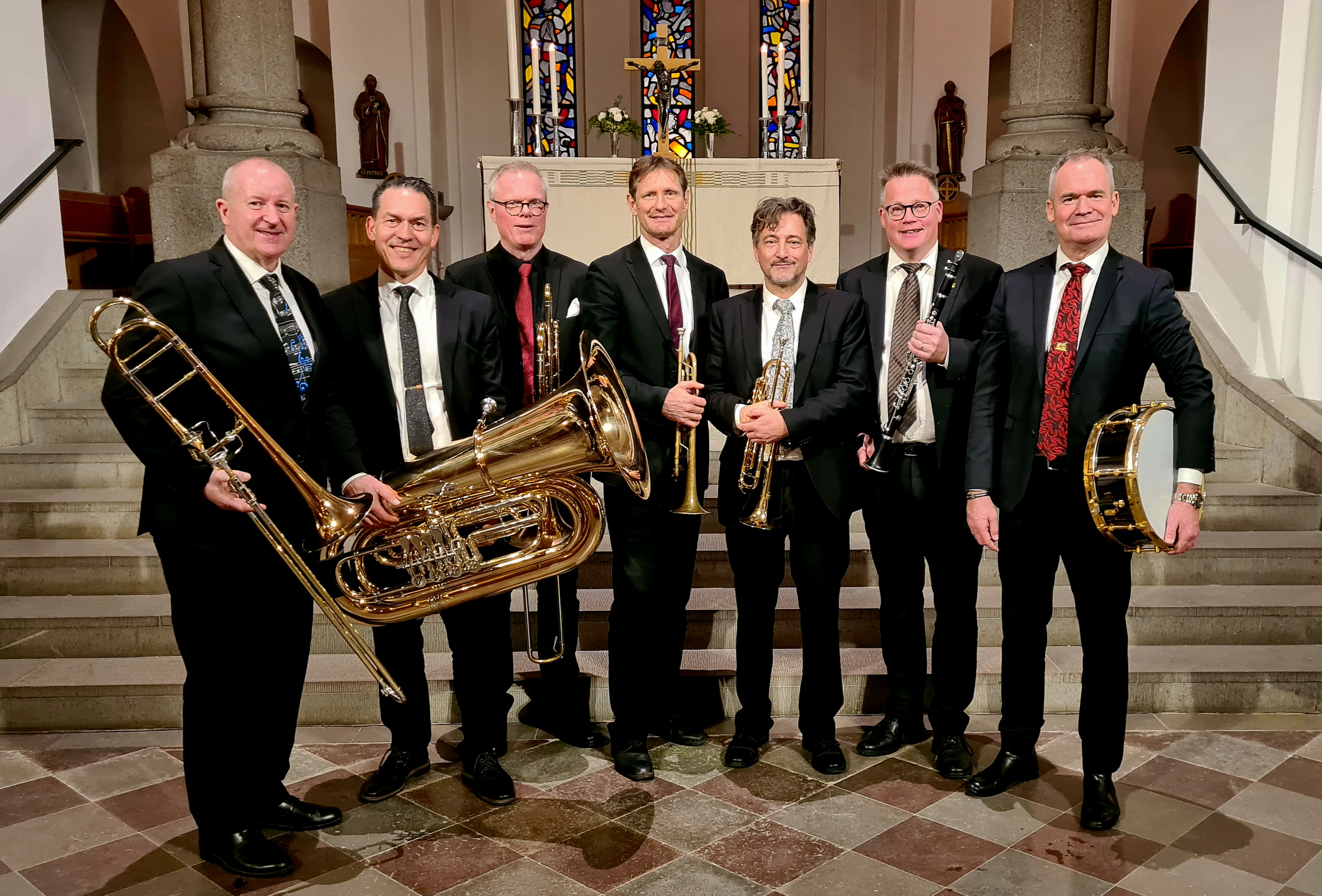 Sju kostymklädda män med blåsinstrument i händerna står på rad framma i koret i Hässleholms kyrka.