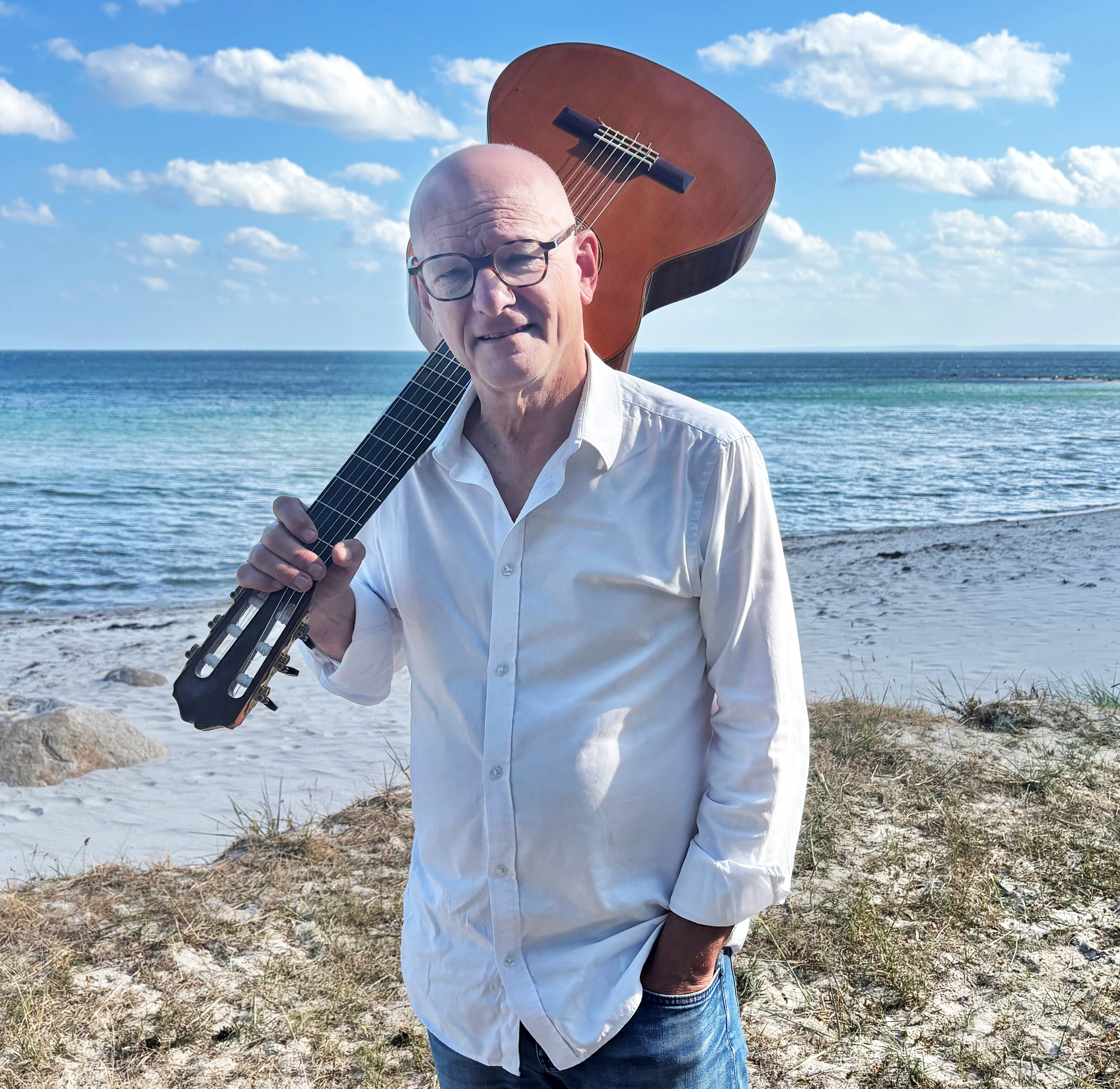 En man med glasögon och vit skjorta går längs med havet med en gitarr över axeln.