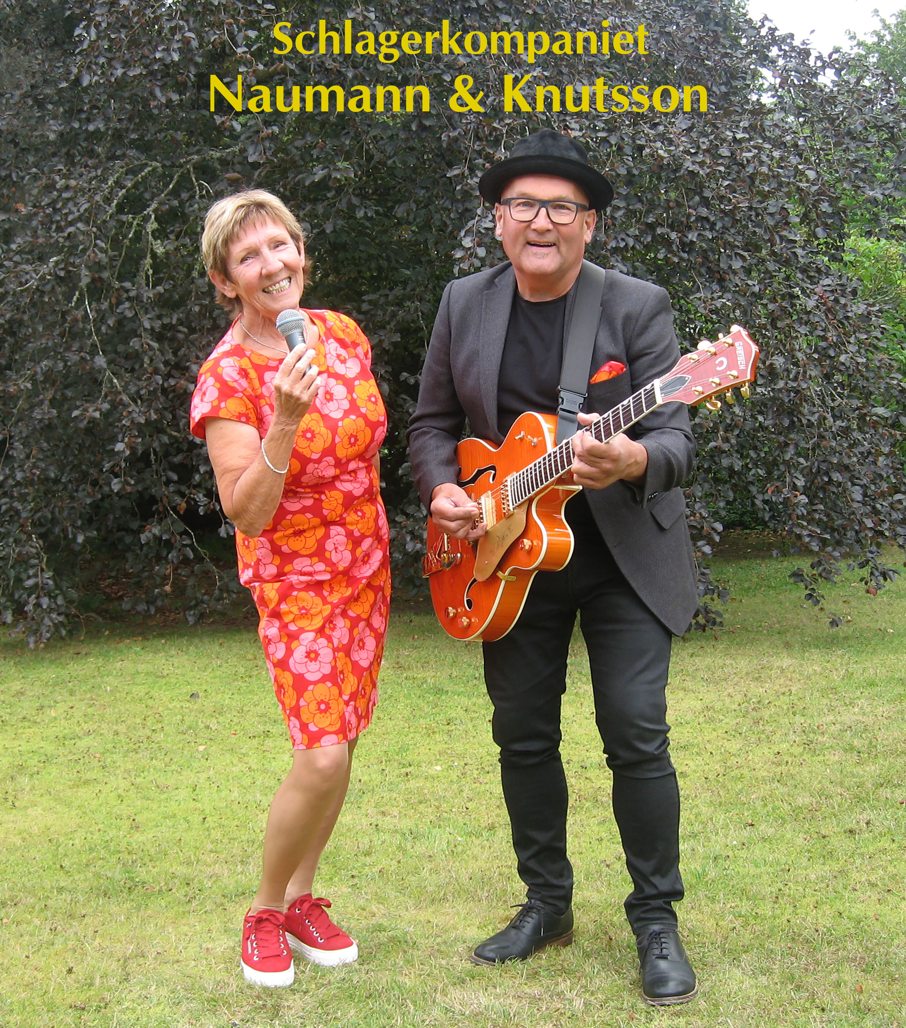 Kvinna med mikrofon och orange klänning och man med svart hatt, gråsvarta kläder och en orange gitarr står på en gräsmatta.