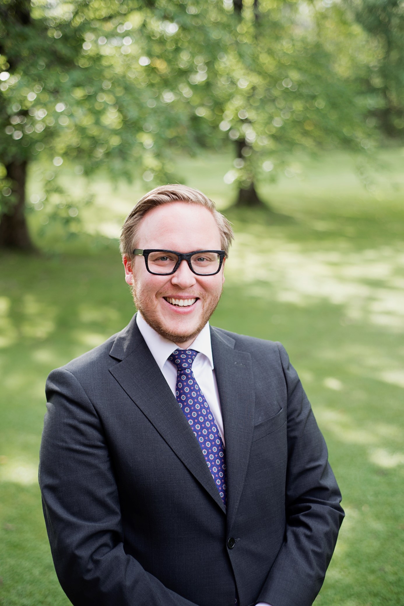 Leende man med glasögon, välklippt skägg, mörk kavaj och blålilamönstrad slips