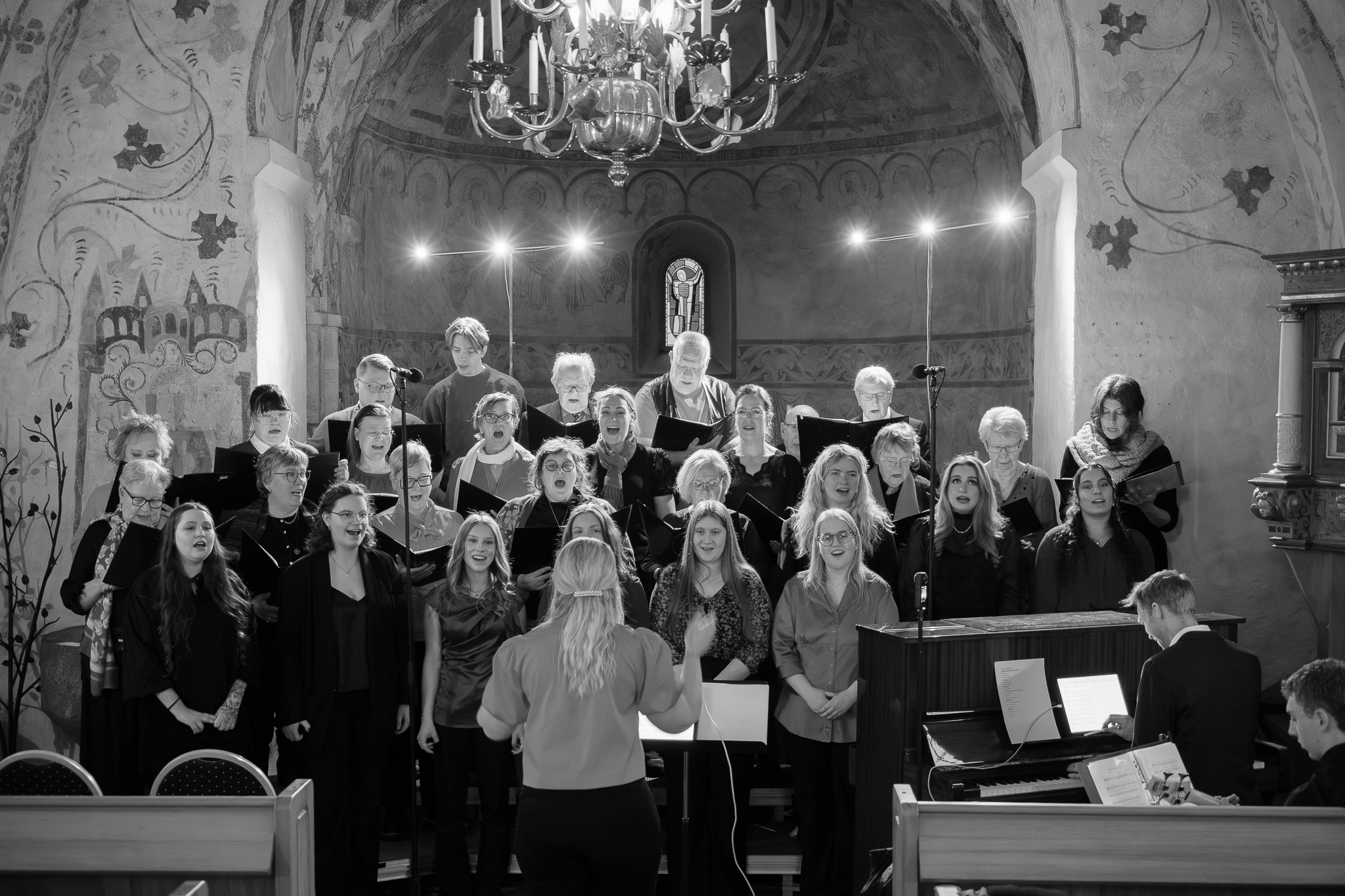 En stor kör står i koret i Hästveda kyrka. Körledaren står med ryggen mot församlingen och dirigerar.