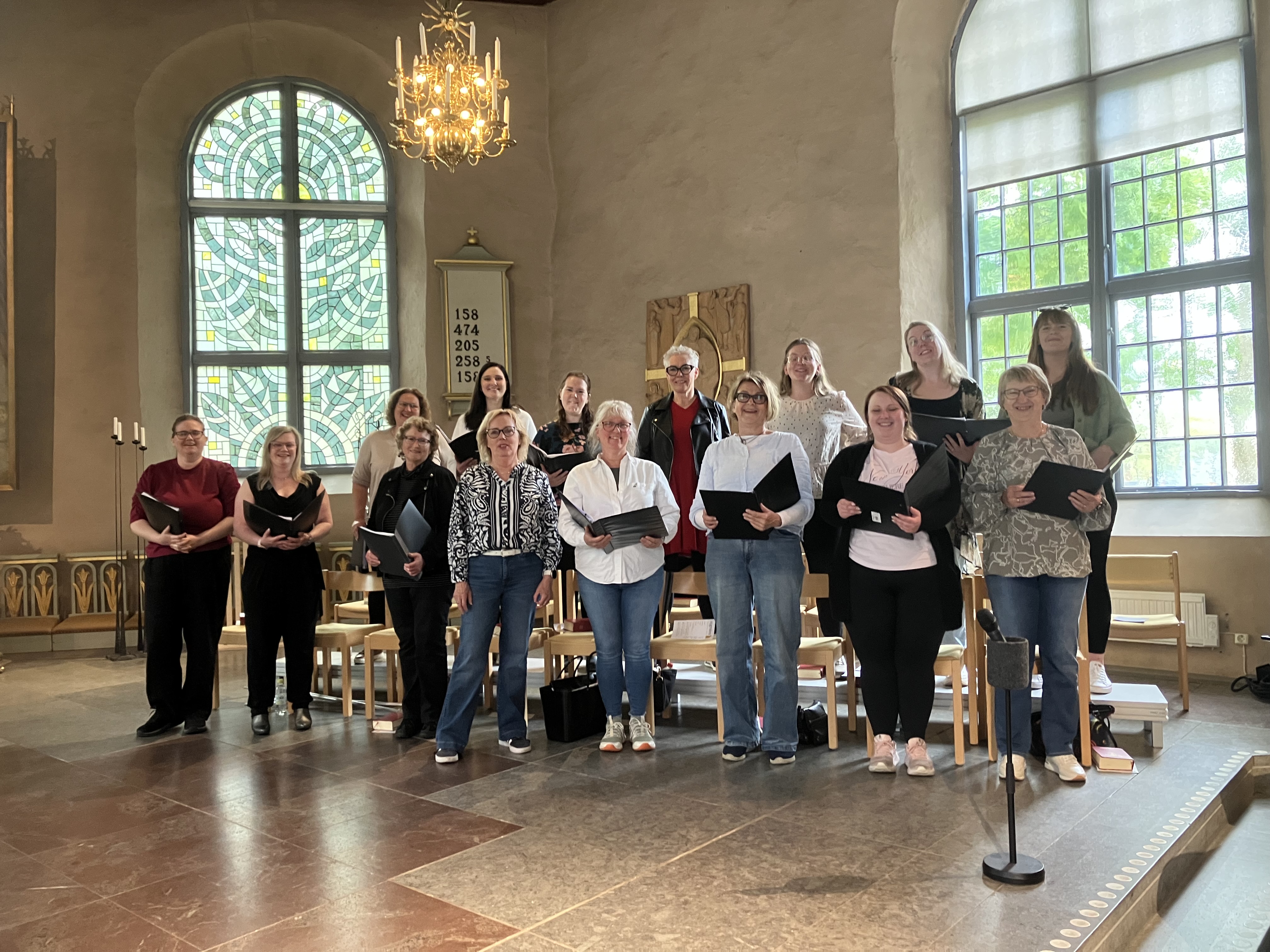 En kör med kvinnor i blandade åldrar sjunger i koret i en kyrka, Christine kyrka, Alingsås, med stora fönster med mycket ljusinsläpp.