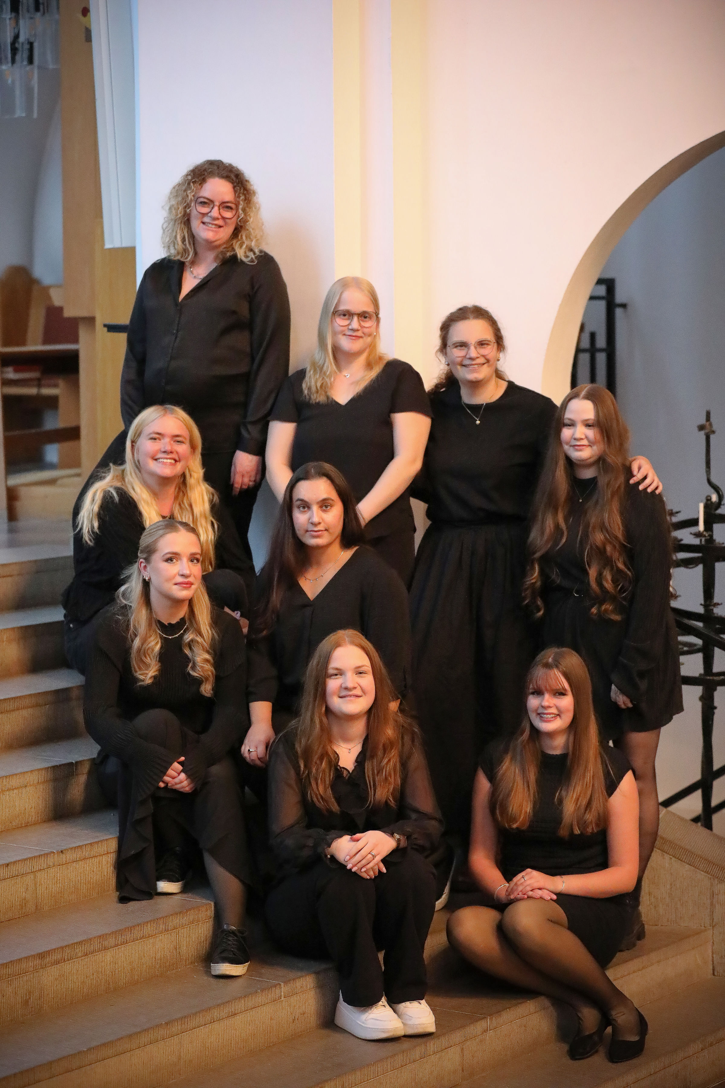 En grupp glada, svartklädda tjejer står i trappan upp till koret i Hässleholms kyrka under en paus i körövningen.