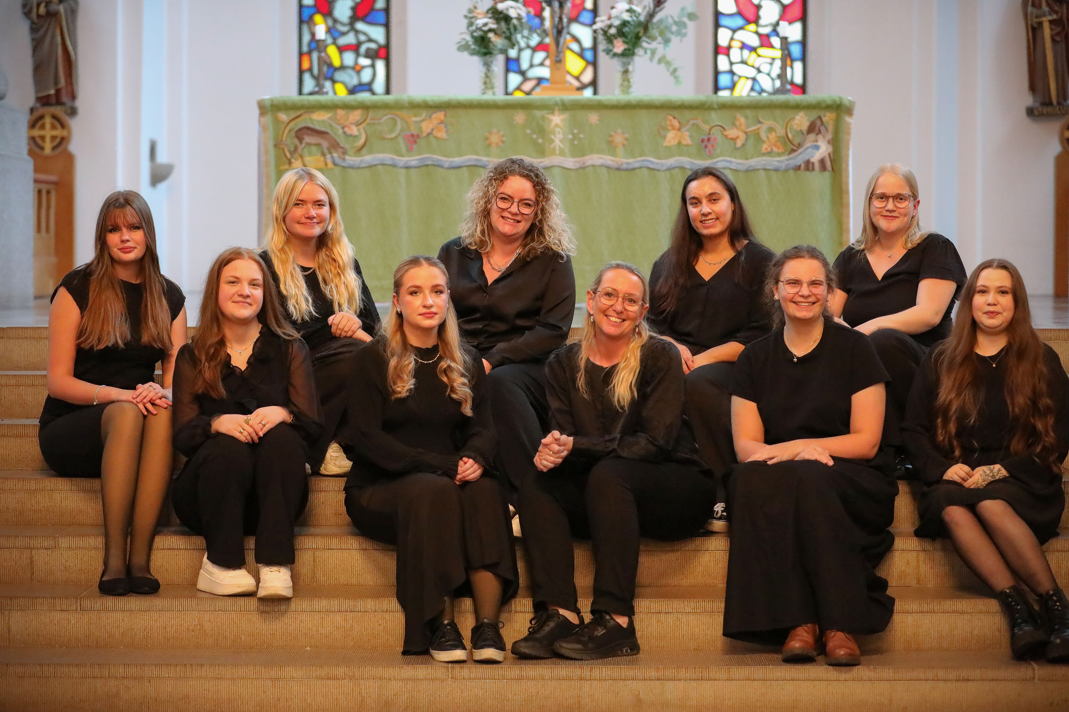 Tio svartklädda tjejer som ser glada ut sitter i trappan till koret i Hässleholms kyrka. I bakgrunden syns altaret med grönt antependium och glasfönsterna i korväggen.