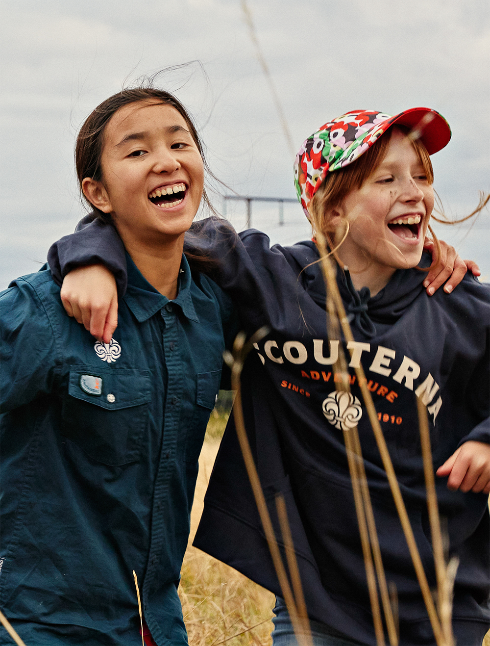 Två glada flickor håller armarna om varandras axlar. De är scouter. Den ena flickan har långt mörkt hår samlat i en hästsvans och den andra flickan har långt brunt hår och en blommig keps.
