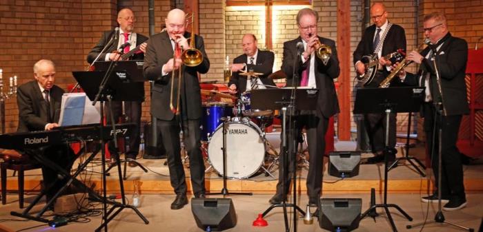 Sju herrar i mörka kostymer spelar instrument i koret i en kyrka. På väggen bakom dem finns ett belyst krucifix.
