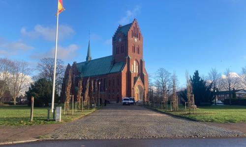 Blå himmel över Hässleholm kyrkas röda tegel. 21 Pelarekar har planterats längs med gången upp mot kyrkan.