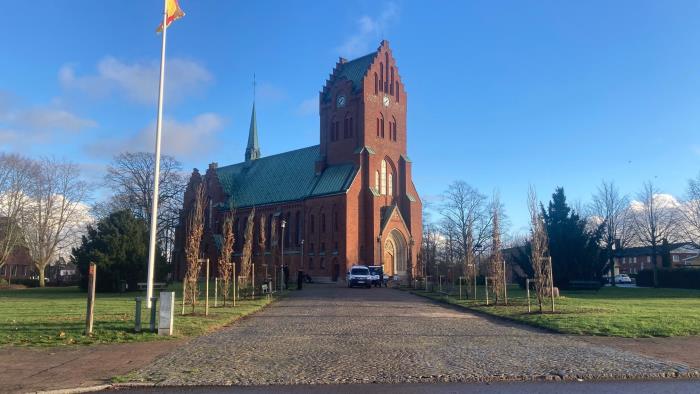 Blå himmel över Hässleholm kyrkas röda tegel. 21 Pelarekar har planterats längs med gången upp mot kyrkan.