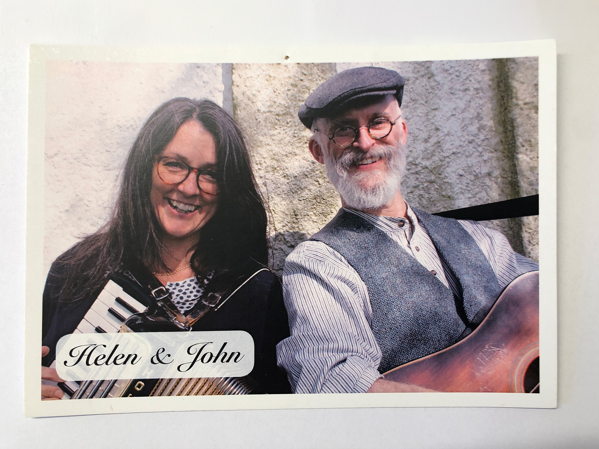 En leende kvinna med långt mörkt hår, glasögon och dragspel och en leende man med grått skägg, mustasch, keps, glasögon och en gitarr.