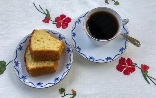 Vit kaffekopp med blått mönster längs kanten. Fat med två skivor sockerkaka. Vit linneduk broderad med röda blommor. Silversked.