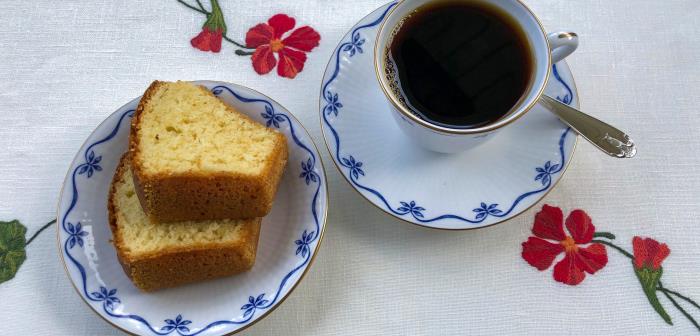 Vit kaffekopp med blått mönster längs kanten. Fat med två skivor sockerkaka. Vit linneduk broderad med röda blommor. Silversked.