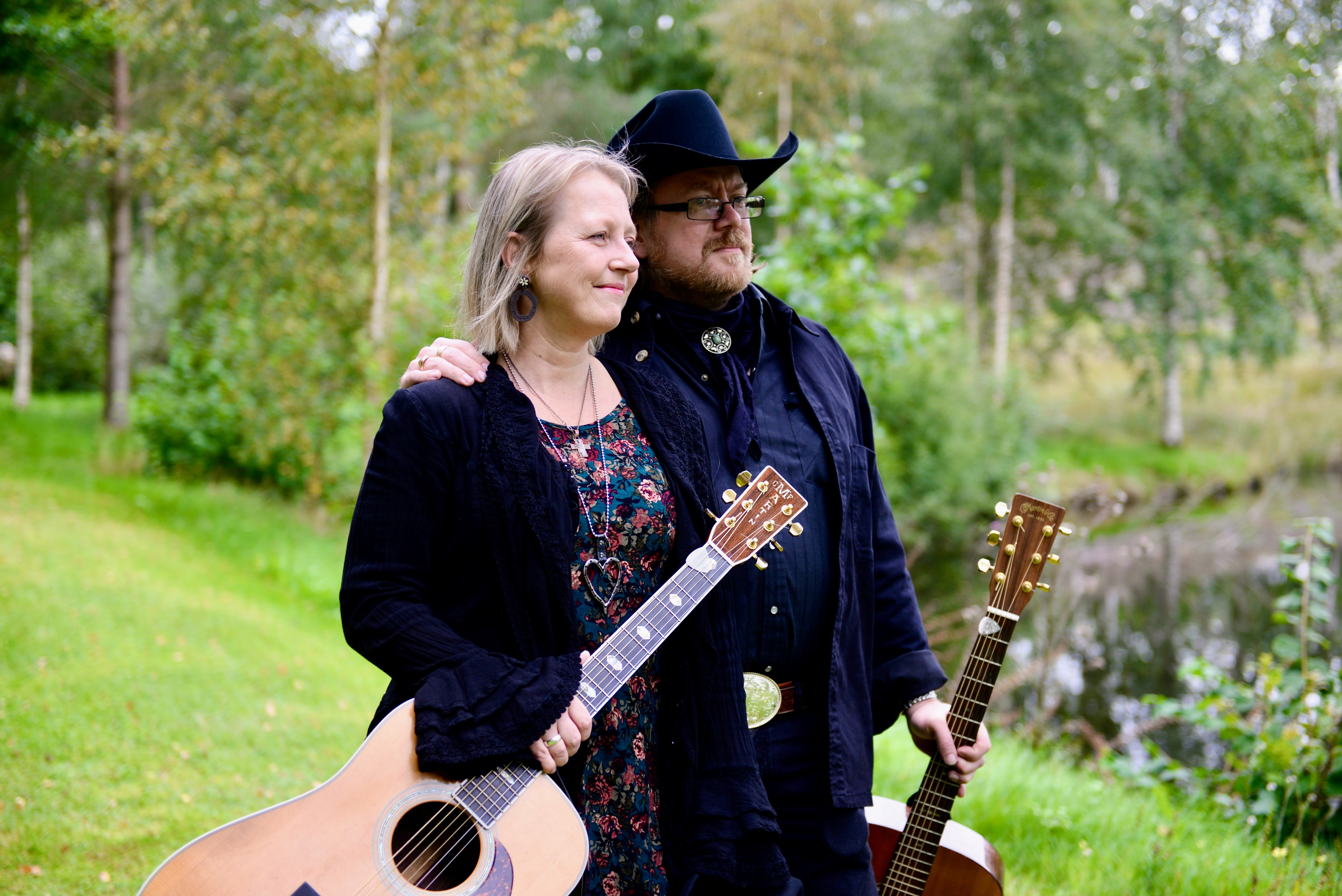 En man med mörka kläder och hatt med uppsvängda bretten och en kvinna med mörka kläder och halvlångt hår. De har varsin gitarr i händerna.
