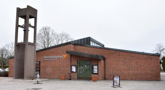 Klockstapel i grå betong framför en låg kyrkobyggnad i rödbrunt tegel.