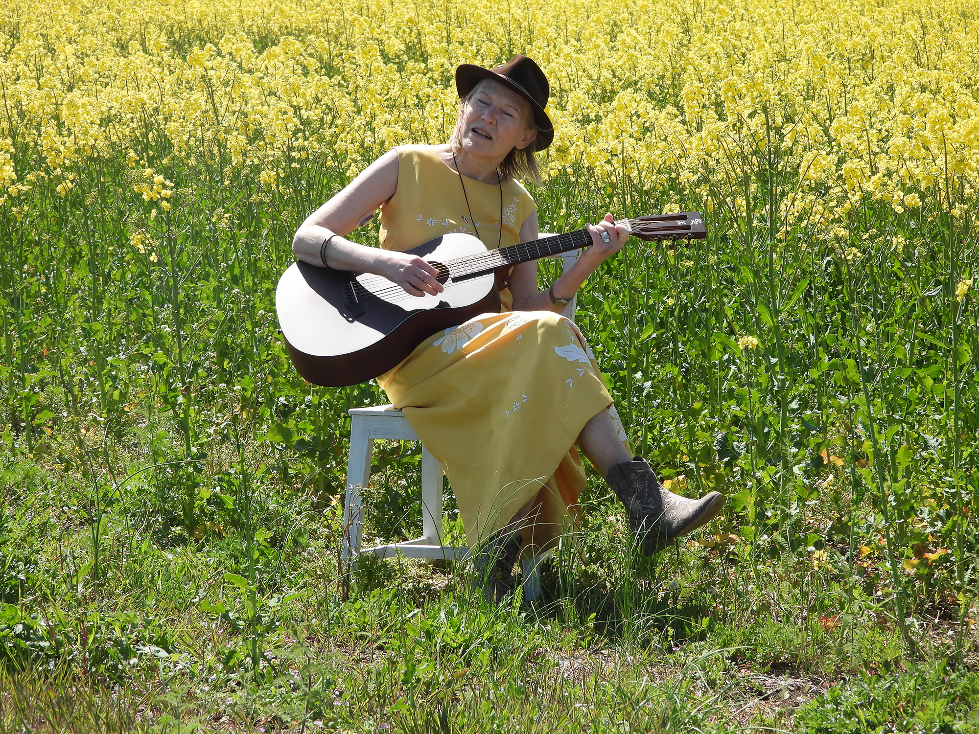 Kvinna i gul klänning och brun hatt sitter på en vit trästol i ett rapsfält och spelar gitarr.